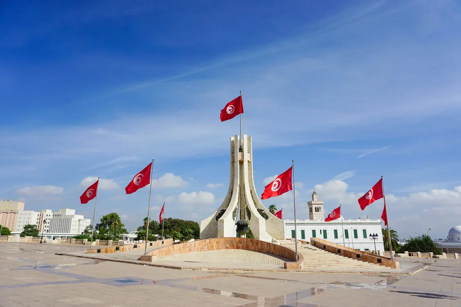 Drapeaux tunisiens flottant au vent sur une place publique, devant un bâtiment historique blanc aux volets verts.